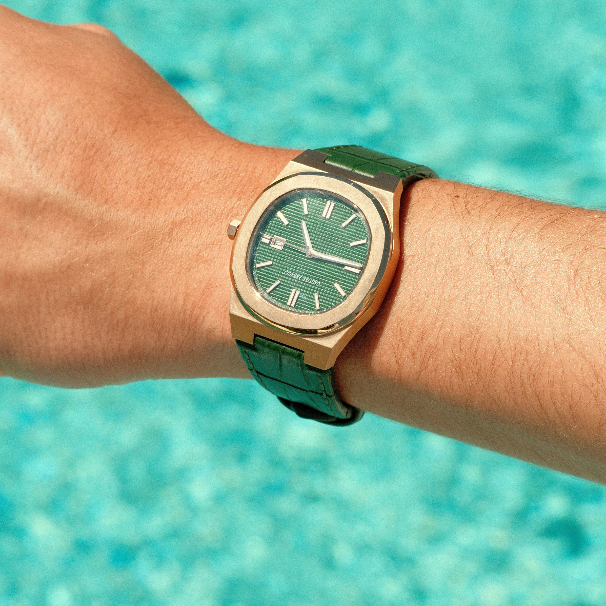 Green watch with gold case on a wrist against a blurred pool background